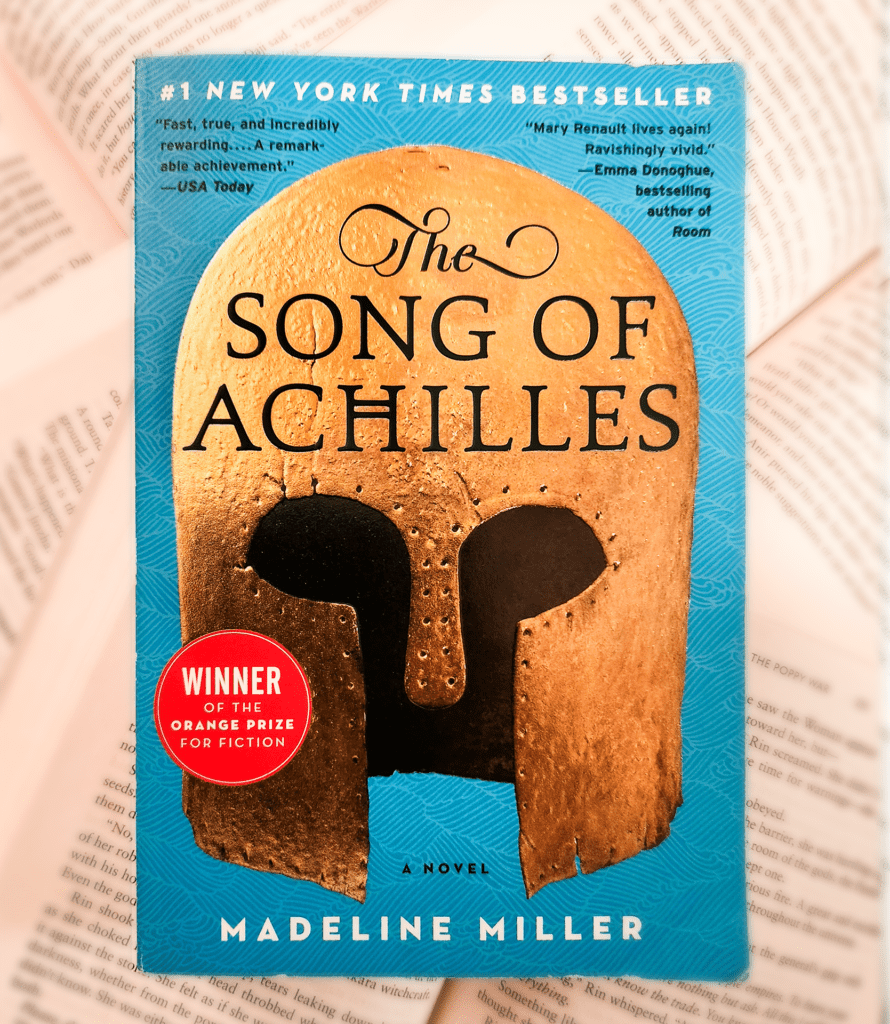 photo of a Ya fantasy book called the song of Achilles by Madeline Miller. closed cover displayed in front of other open books on the background.